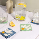 Punch needle coaster in blue and mint green, lemon water in a jug with glasses atop the coasters. A slice of lemon lies in the foreground.