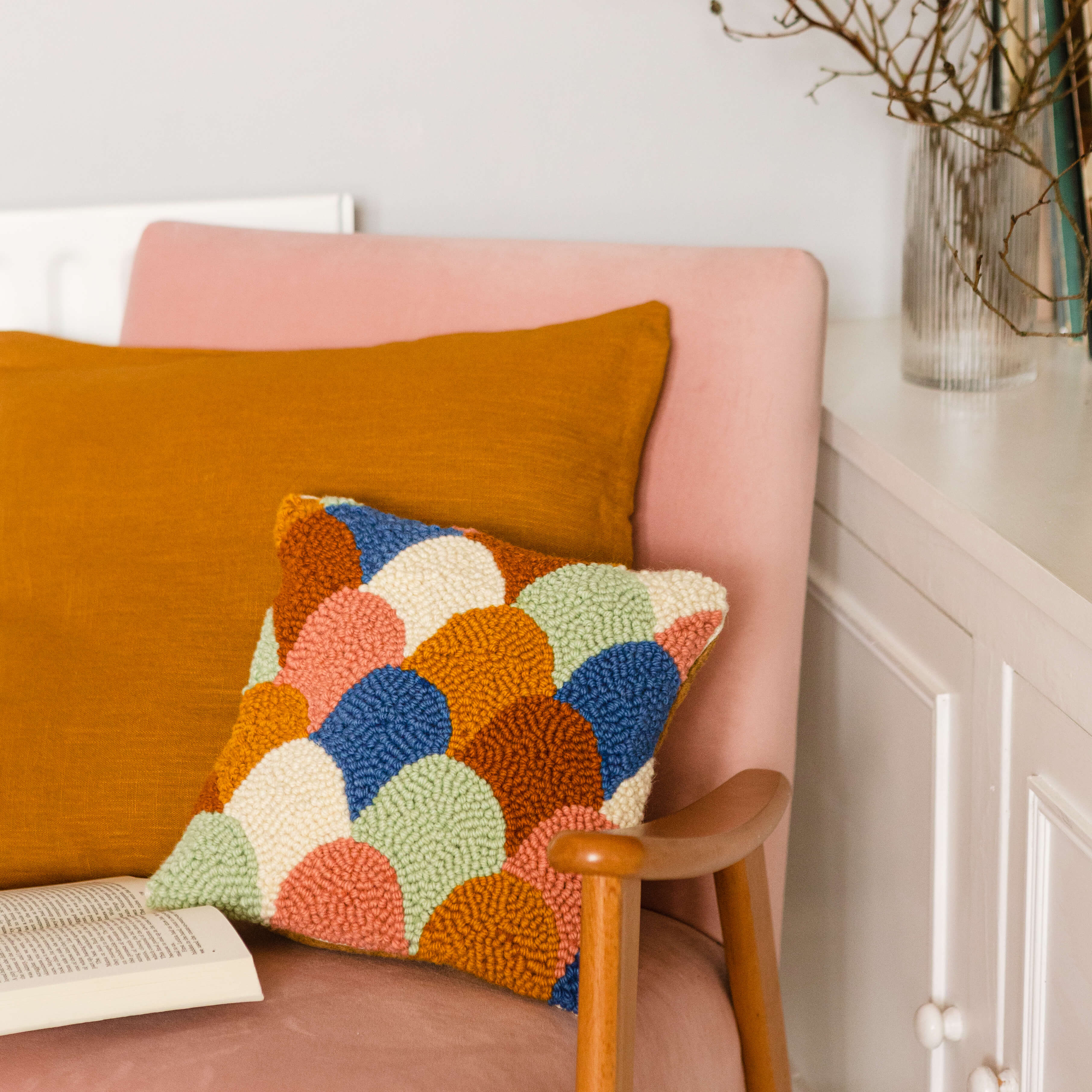 Scallop punch needle cushion propped on light pink chair with tan cushion and book open